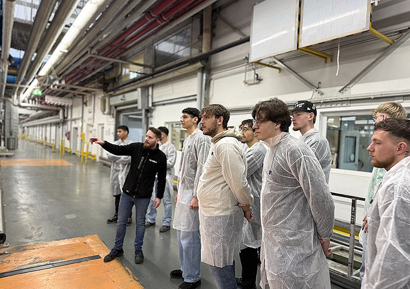 Bei der Begehung macht Damian auf das führerlose Transportsystem im Pappenwerk aufmerksam. Schüler folgen aufmerksam.
