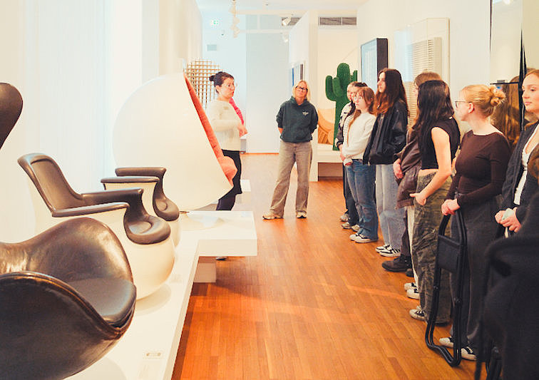 Eine Gruppe von Besucherinnen und Besuchern steht in einer Ausstellung des Museum für Angewandte Kunst Köln mit futuristischen Sitzmöbeln und Designobjekten. Die Museumsführerin erklärt die ausgestellten Werke.