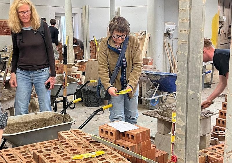 Zwei Lehrerinnen stehen in einer Maurerwerkstatt inmitten von Ziegelsteinen