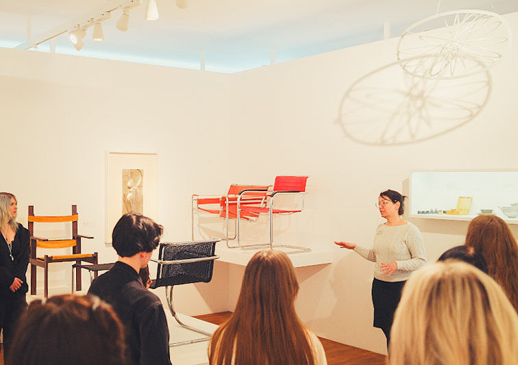 Besuchergruppe hört einer Führung in der Designausstellung des Museum für Angewandte Kunst Köln zu. Im Raum befinden sich moderne Möbel und Designklassiker.