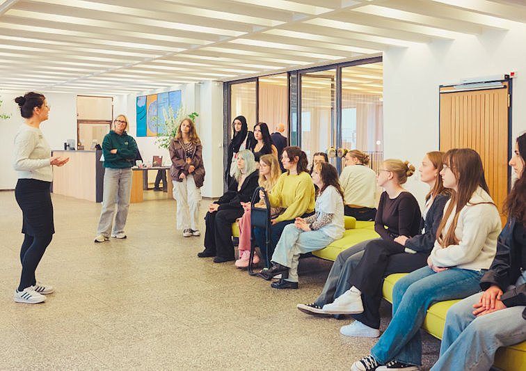 Eine Museumsführerin spricht in der Eingangshalle des Museum für Angewandte Kunst Köln zu einer Gruppe junger Besucher/-innen. Einige sitzen auf einer grünen Bank, andere stehen im hellen, modern gestalteten Raum.