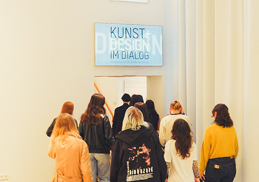 Eine Besuchergruppe geht durch einen breiten Flur des Museum für Angewandte Kunst Köln in Richtung eines Ausstellungsbereichs. Über dem Eingang hängt ein Schild mit der Aufschrift „Kunst + Design im Dialog“.