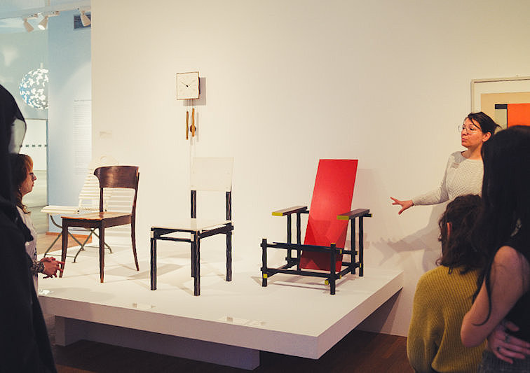 Eine Museumsführerin erklärt Designstühle und Möbelobjekte in einer Ausstellung des Museum für Angewandte Kunst Köln. Die Objekte stehen auf weißen Podesten in einem hellen Raum.